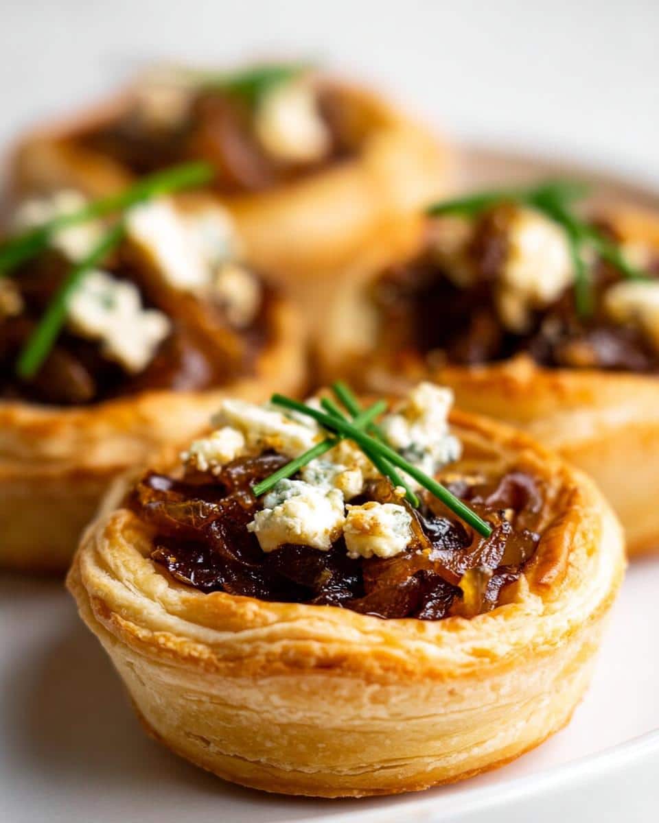 A close-up of a flaky puff pastry Caramelized Onion Tartlet topped with dark, rich onions, crumbled blue cheese, and fresh chives.