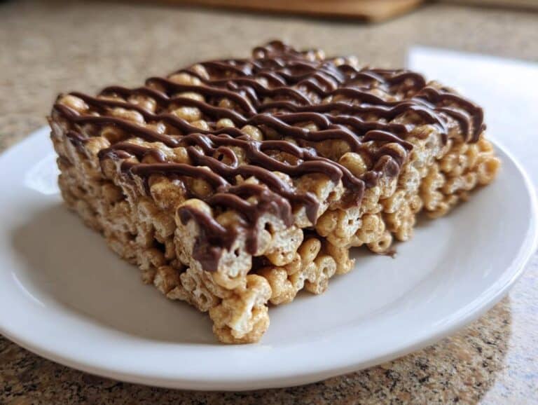 A close-up of a square Cheerio Marshmallow Snack Bars piece drizzled with melted chocolate on a white plate.