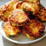 A pile of golden brown, crispy Parmesan-Crusted Zucchini Bites stacked on a white plate.