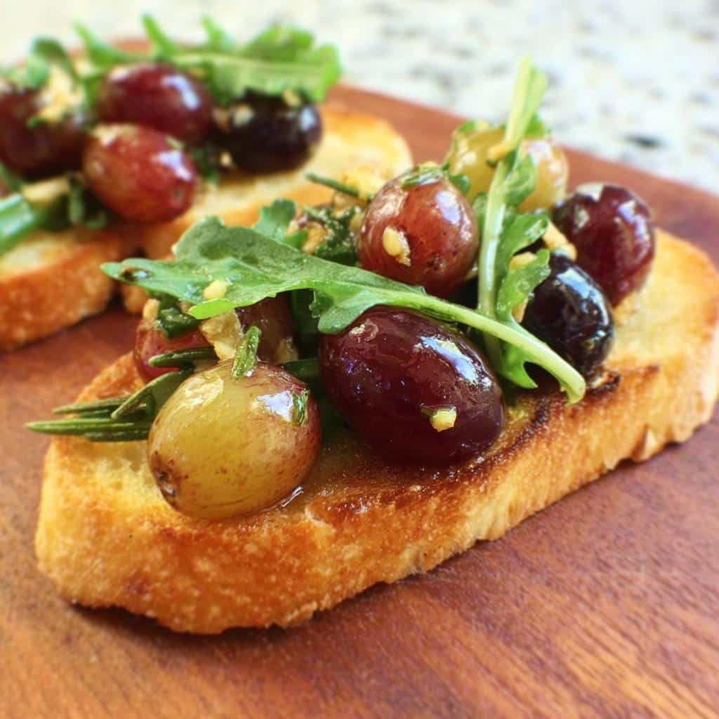 Close-up of a toasted baguette slice topped with roasted grapes, rosemary, and arugula for Roasted Grape & Rosemary Crostini.