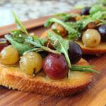Close-up of a single Roasted Grape & Rosemary Crostini topped with roasted grapes, arugula, and rosemary on a wooden board.