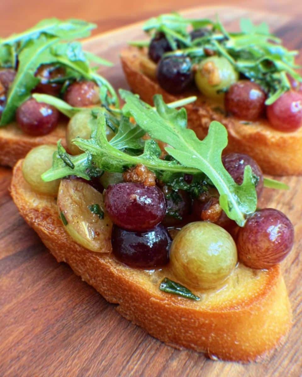 Close-up of Roasted Grape & Rosemary Crostini topped with fresh arugula on a wooden board.