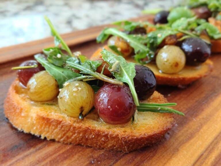 Close-up of a single Roasted Grape & Rosemary Crostini topped with roasted grapes, arugula, and rosemary on a wooden board.