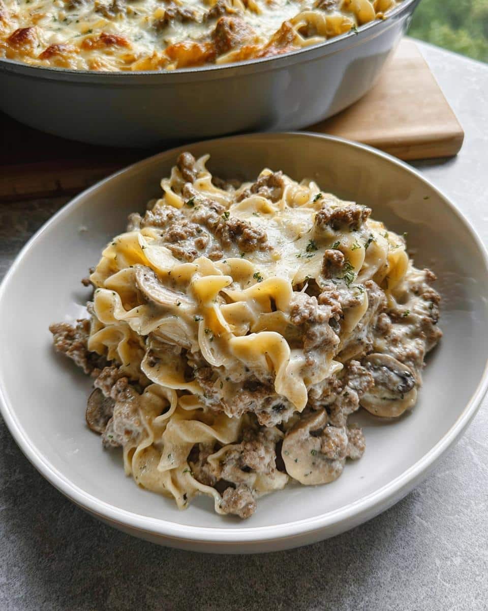 A bowl filled with creamy beef and mushroom casserole mixed with egg noodles, with the main baking dish visible in the background.
