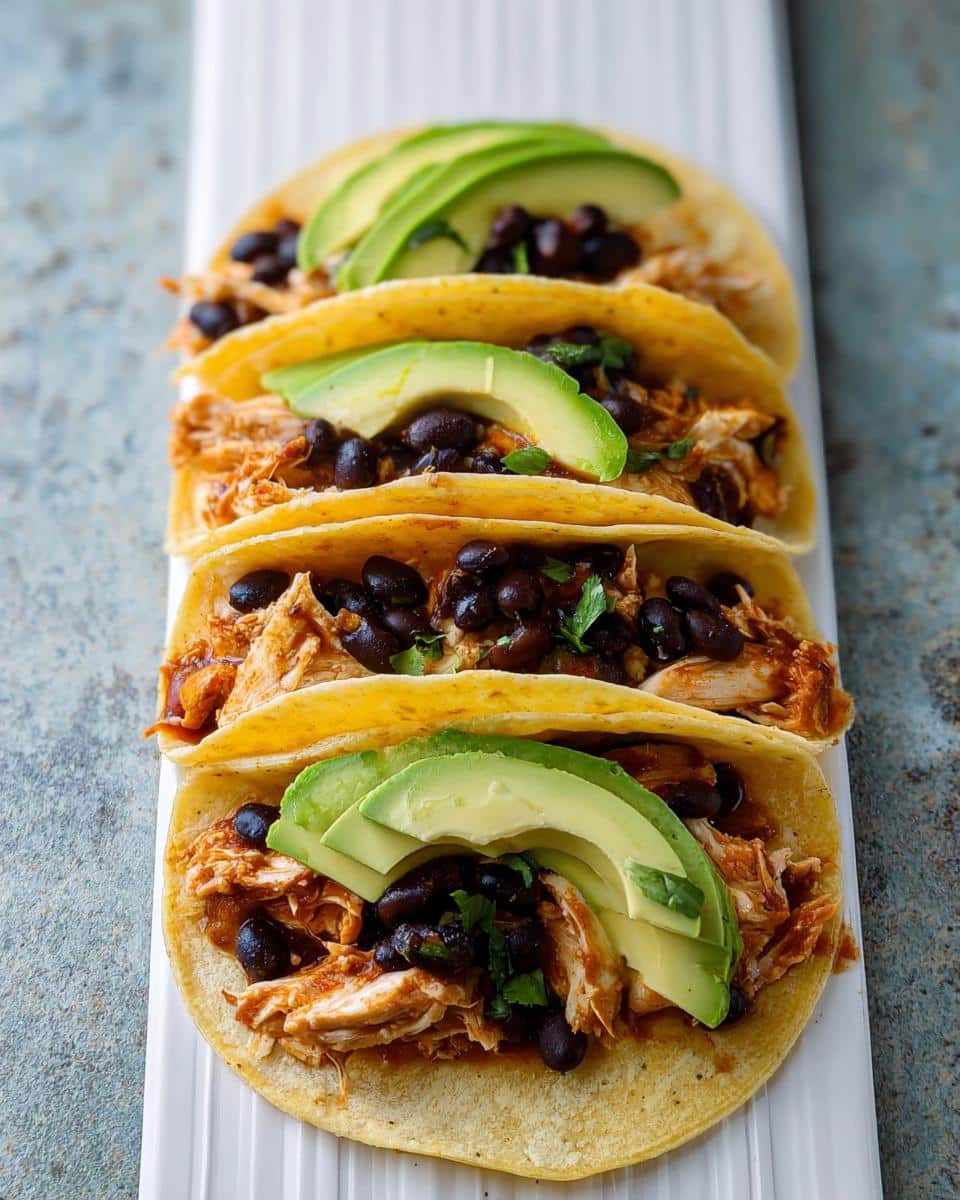 Four soft corn tortillas filled with shredded chicken, black beans, and topped with sliced avocado, ready to eat.