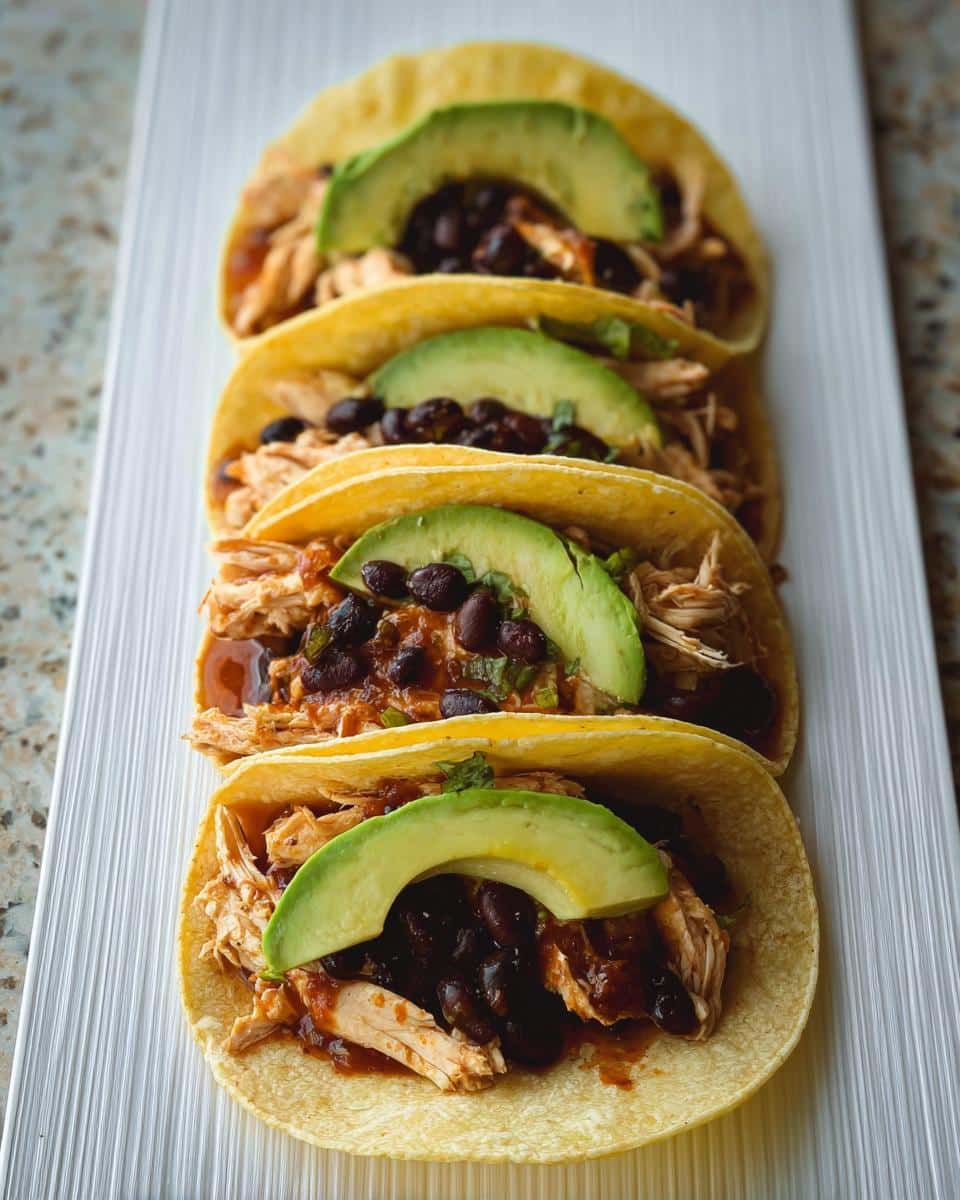 Four corn tortillas filled with shredded chicken, black beans, and topped with avocado slices for 5-Ingredient Chicken Tacos.