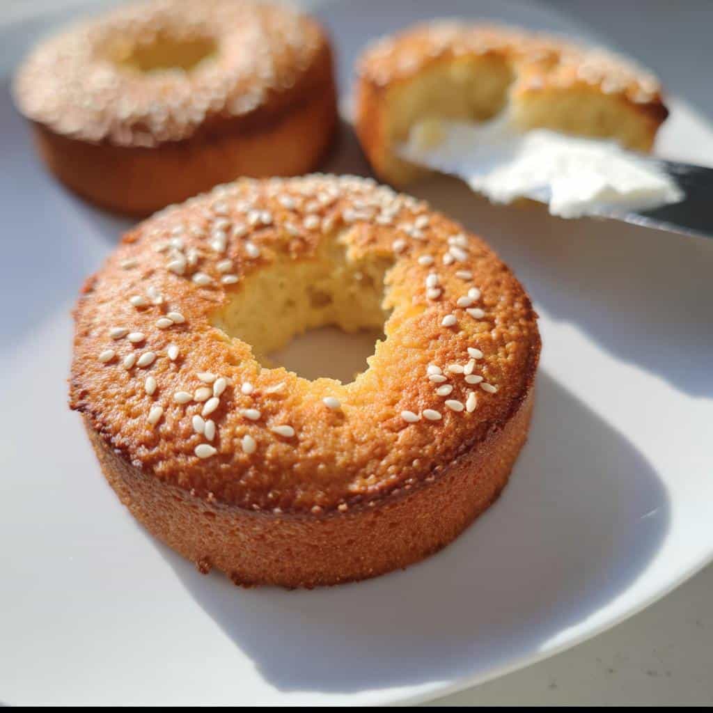 Close-up of golden Keto Coconut Flour Bagels topped with sesame seeds on a white plate, one being spread with cream cheese.