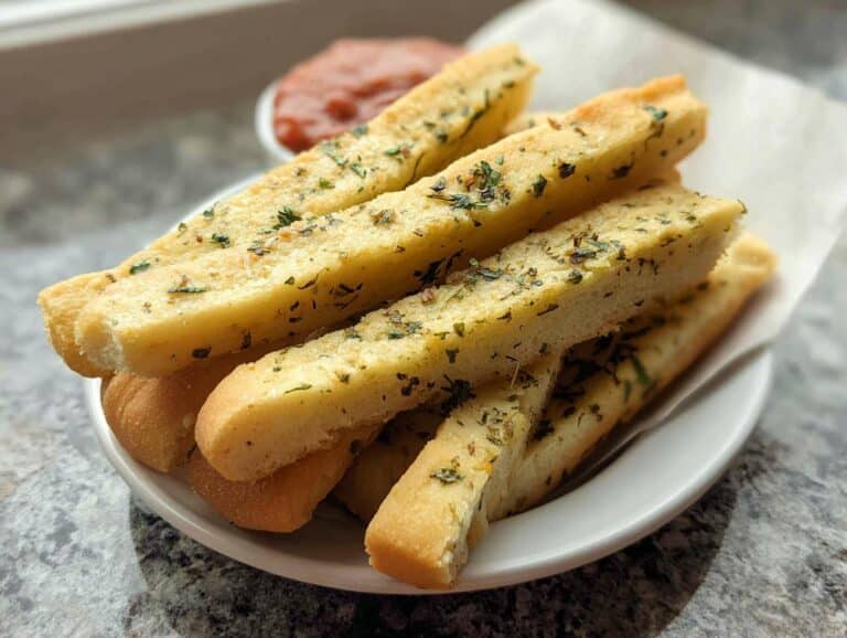 A stack of golden No-Yeast Herb Breadsticks seasoned with dried green herbs, served on a white plate with dipping sauce in the background.