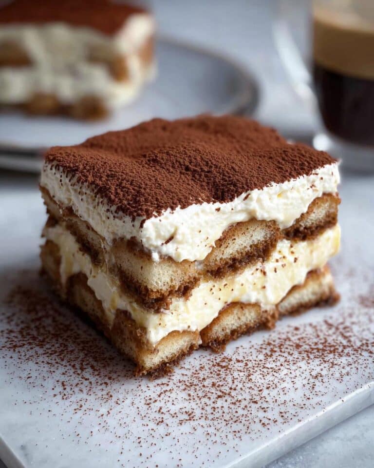 A perfect square slice of Quick Thermomix Tiramisu featuring layers of cream and coffee-soaked ladyfingers, topped with cocoa powder.