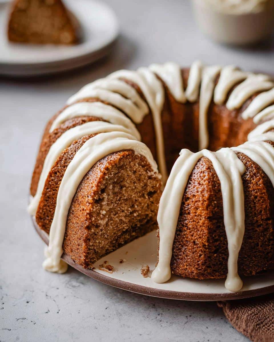 A close-up of a rich Banana Bread Bundt Cake with thick, white cream cheese frosting drizzled over the top, showing a slice removed.
