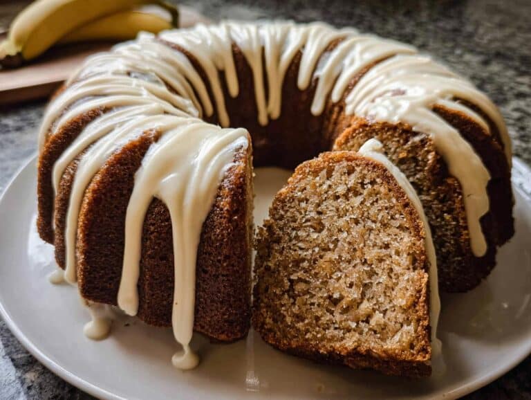 A rich Banana Bread Bundt Cake, drizzled with white glaze, with one slice cut out showing the moist interior.