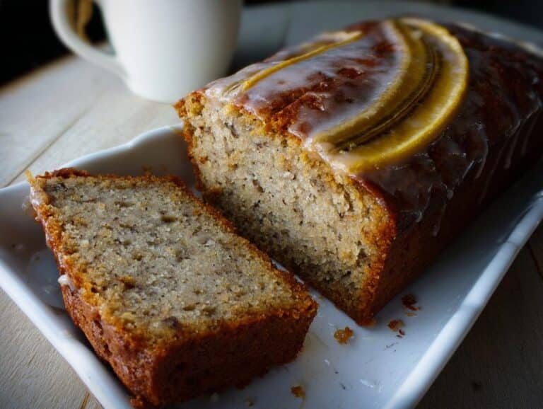 A loaf of moist Banana Bread Citron, partially sliced, topped with a lemon glaze and banana slices.