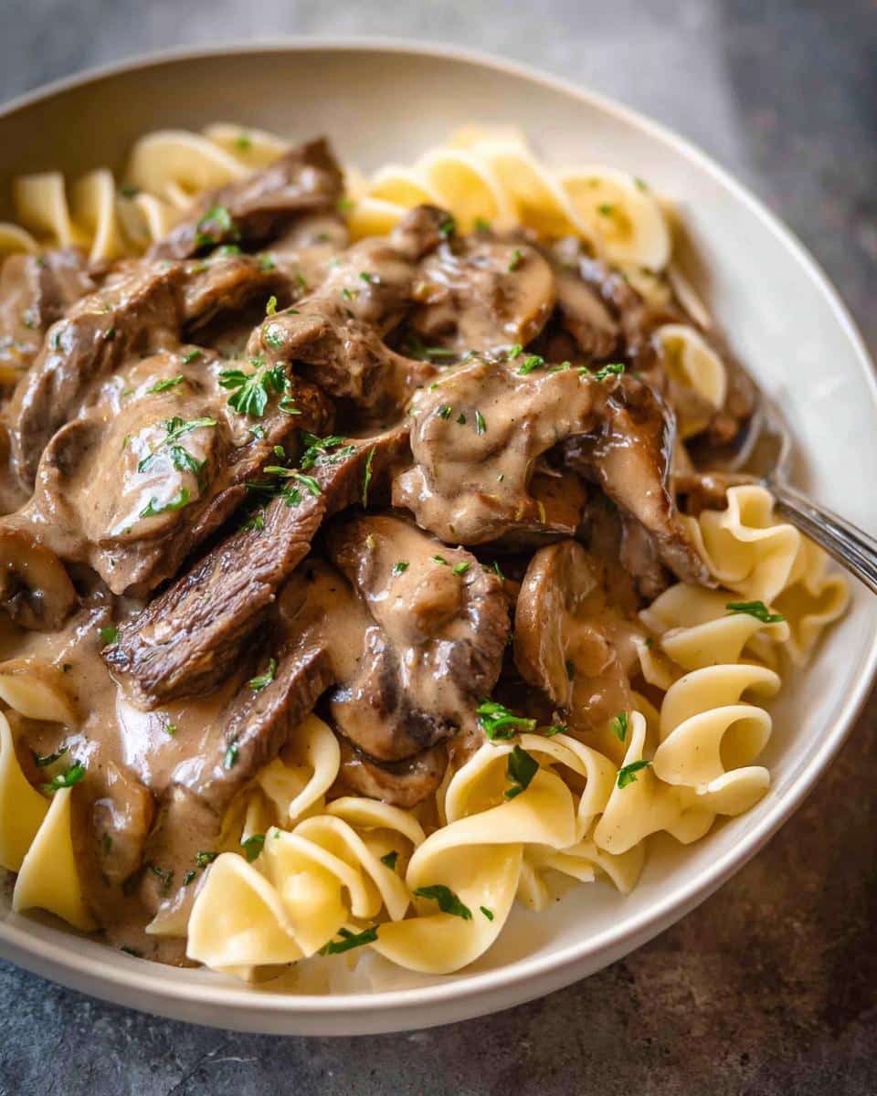Close-up of a bowl featuring creamy Beef Stroganoff Dinner with sliced beef and mushrooms over egg noodles, garnished with parsley.