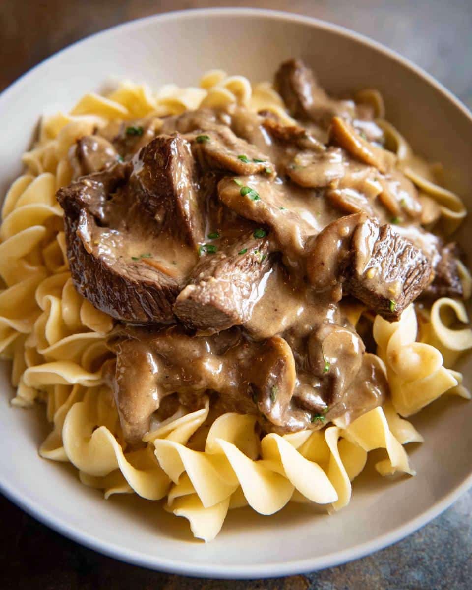 A close-up of a hearty Beef Stroganoff Dinner featuring tender beef and mushrooms in a creamy sauce served over wide egg noodles.