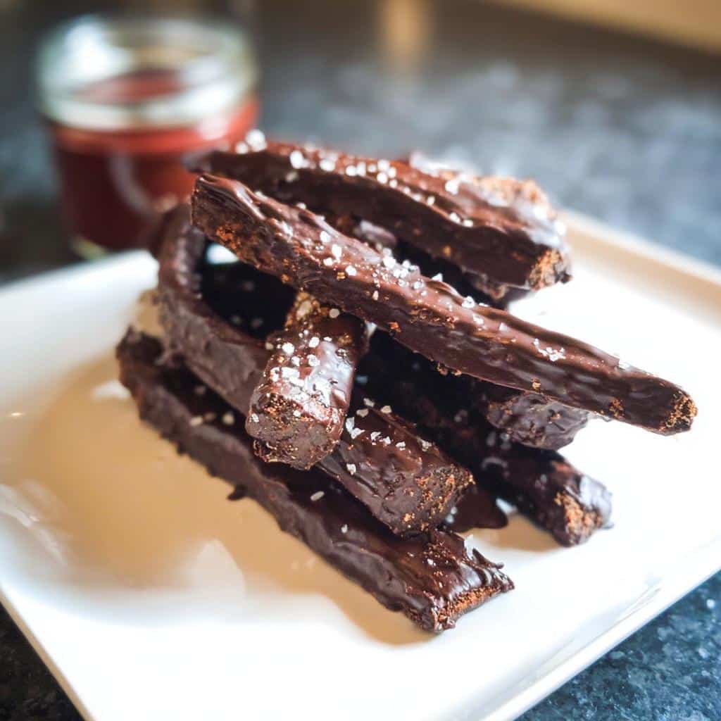 A stack of rich, chocolate-dipped Brownie French Fries sprinkled with flaky sea salt on a white plate.