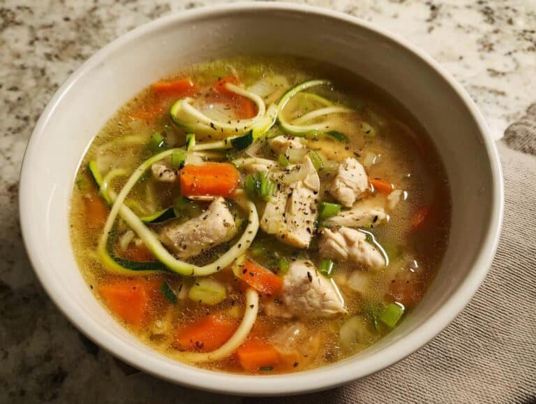 Close-up of a white bowl filled with Chicken Zoodle Detox Soup, featuring chicken chunks, zucchini noodles, carrots, and broth.