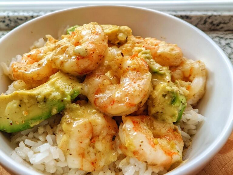 Close-up of a white bowl filled with white rice topped with creamy sauce, seasoned shrimp, and chunks of avocado in a Cold Shrimp Avocado Rice Bowl.