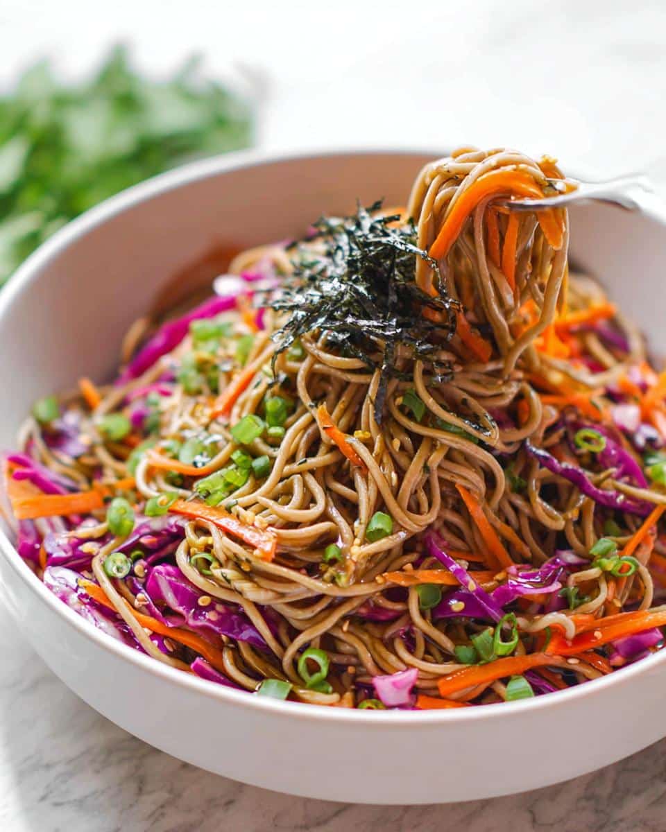 A fork lifts noodles from a vibrant Cold Soba Noodle Lunch Bowl topped with shredded cabbage, carrots, and nori.