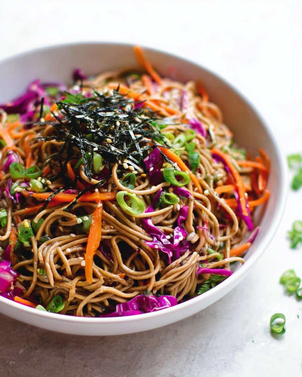 Close-up of a white bowl filled with Cold Soba Noodle Lunch Bowl, topped with shredded carrots, purple cabbage, and nori.