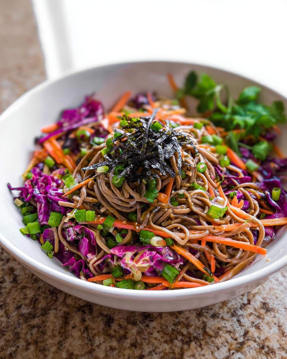 A white bowl filled with Cold Soba Noodle Lunch Bowl, topped with shredded purple cabbage, carrots, and nori.