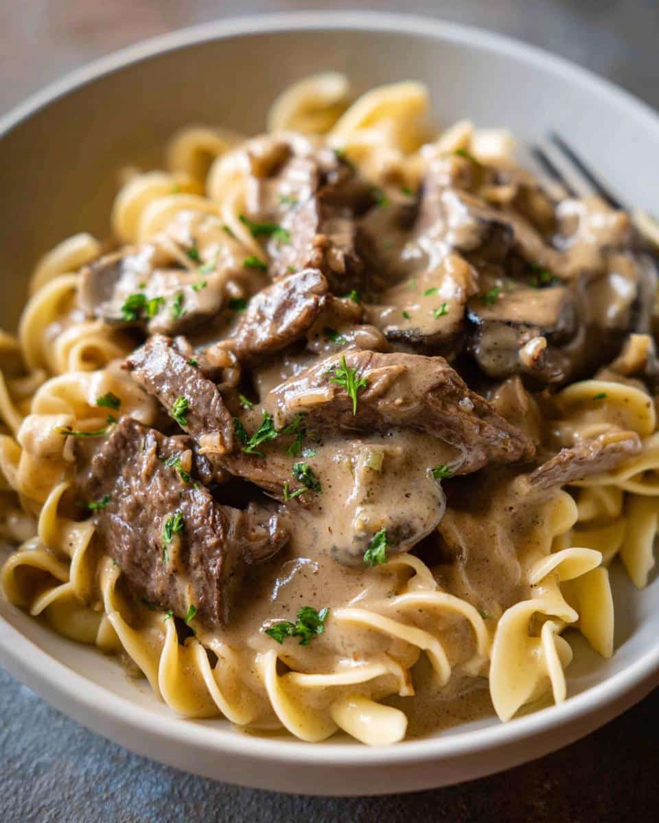 Close-up of a bowl of creamy Beef Stroganoff Dinner with tender beef slices and mushrooms over egg noodles, garnished with parsley.