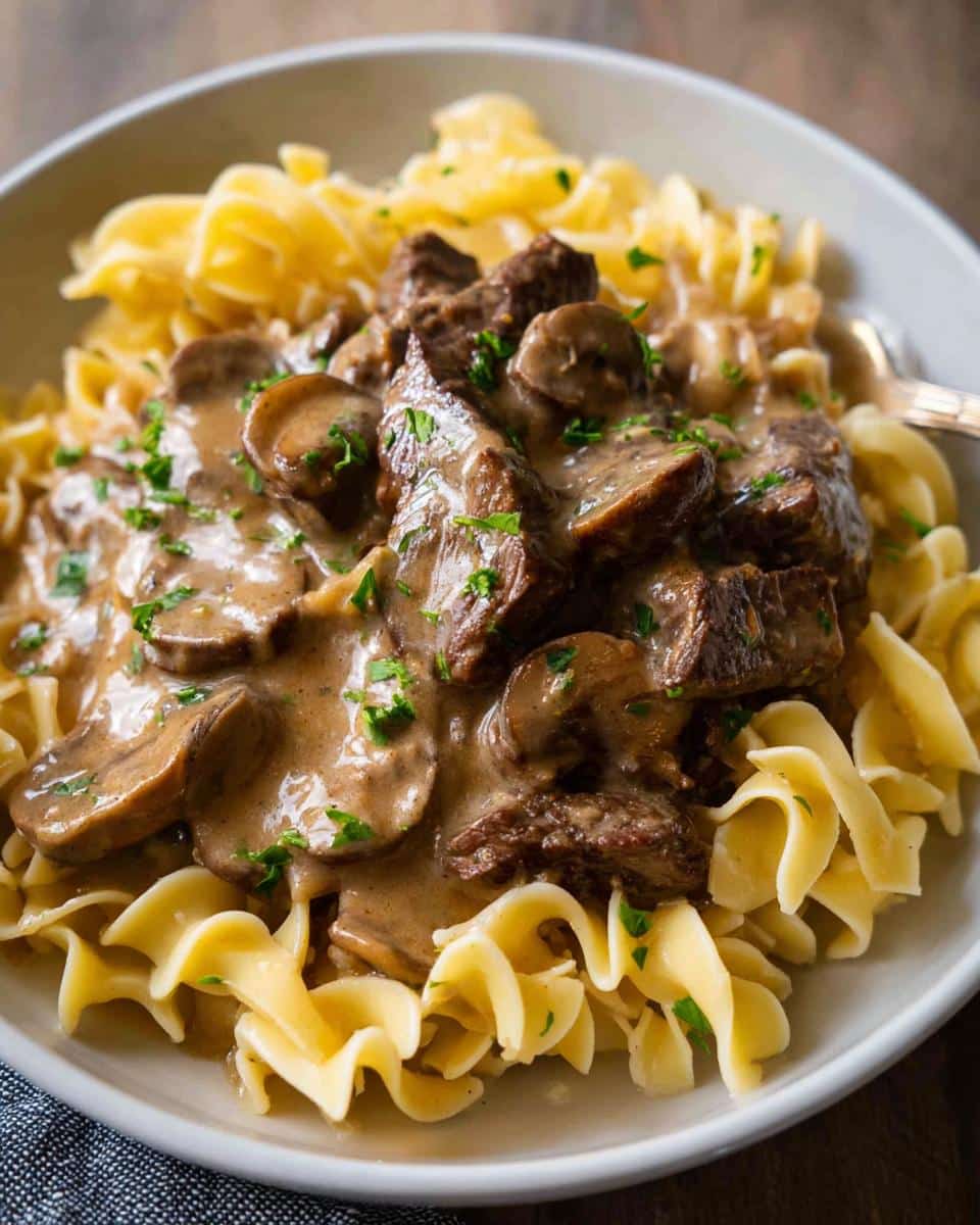 A close-up of a bowl featuring rich Beef Stroganoff Dinner with beef chunks and mushrooms over wide egg noodles, garnished with parsley.