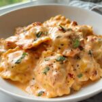 A close-up view of ravioli smothered in a rich, orange-hued creamy sauce, garnished with fresh herbs, part of a Creamy Ravioli Dinner.