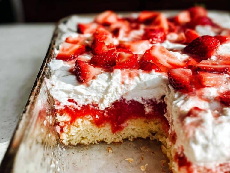 A close-up view of a section missing from an Easy Strawberry Poke Cake topped with whipped cream and fresh strawberries.