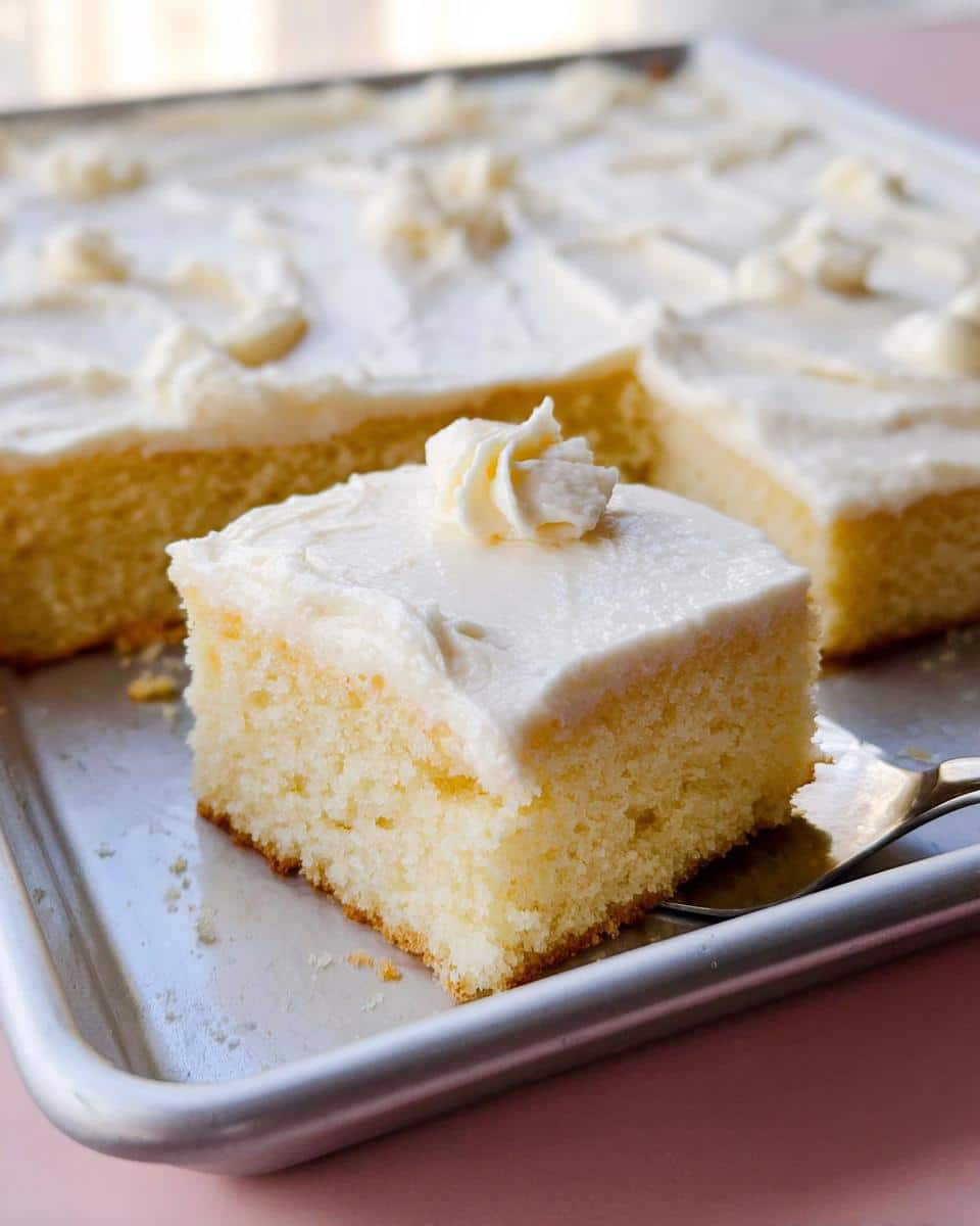 A close-up of a square slice of Easy Vanilla Sheet Cake topped with white frosting, resting on a baking sheet.