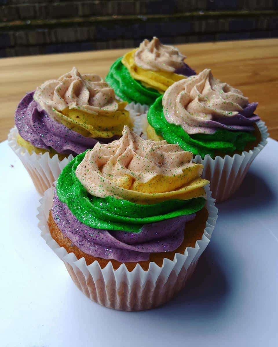 Four Fat Tuesday Dessert cupcakes featuring purple, green, and yellow frosting layers, topped with light pink swirls and glitter.