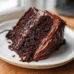 A close-up of a moist slice of rich Gluten Free Chocolate Cake covered in thick chocolate frosting on a white plate.