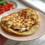 A golden-brown Gluten Free Cottage Cheese Flatbread folded over and stuffed with white meat chicken pieces, served on a pink plate.