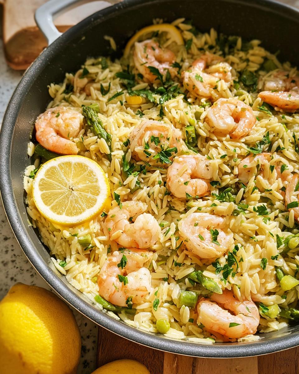 Close-up of Gluten Free Lemon Orzo with Shrimp, asparagus, and lemon slices in a dark skillet.