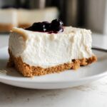 A thick slice of creamy Gluten Free No Bake Cheesecake topped with dark berry compote on a white plate.