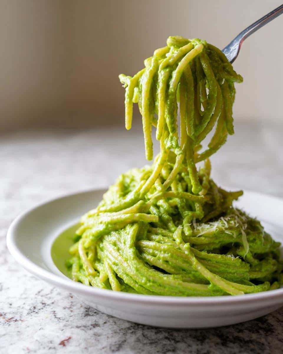 A fork lifting strands of creamy Green Pasta Alfredo from a white bowl sitting on a speckled counter.