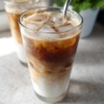 Close-up of a tall, sweating glass filled with a layered Iced White Chocolate Mocha, ice cubes, and milk.