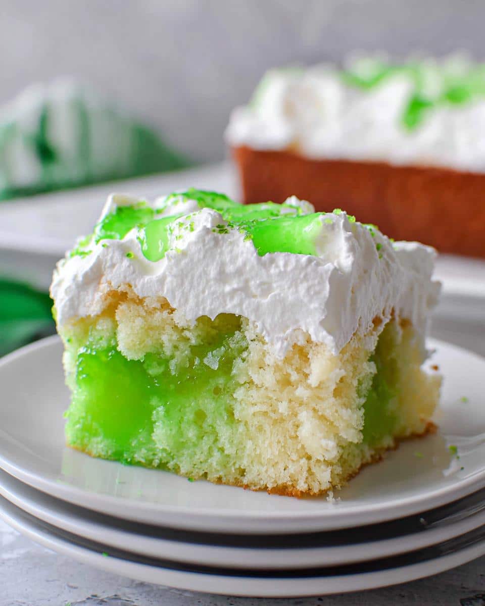 Close-up of a moist slice of Lime Poke Cake showing the bright green gelatin filling absorbed into the white cake, topped with whipped cream.