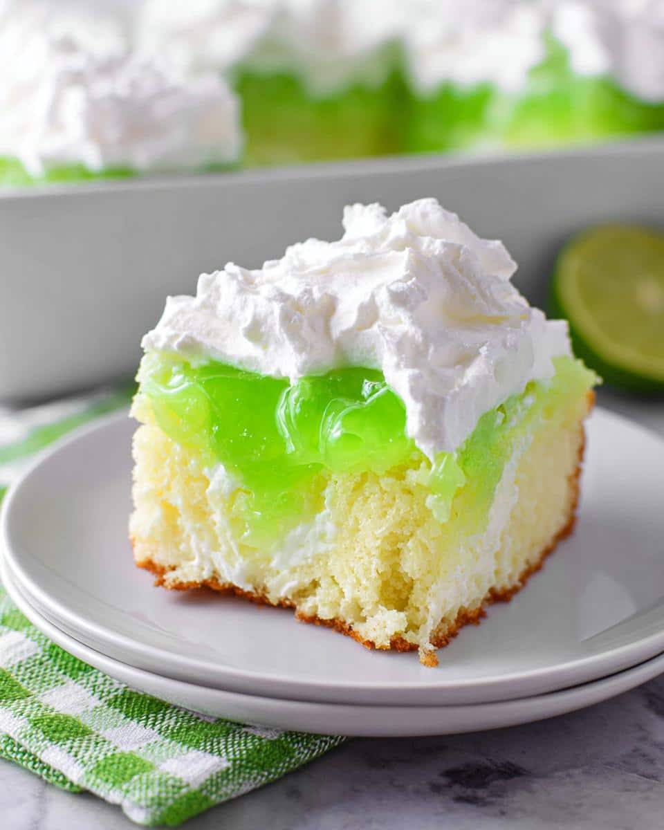 A moist slice of Lime Poke Cake featuring a yellow cake base, white creamy layer, bright green gelatin, and topped with whipped cream.