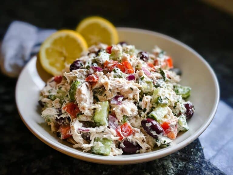 A bowl filled with creamy Low Carb Greek Chicken Salad featuring shredded chicken, cucumbers, tomatoes, and olives, garnished with lemon wedges.