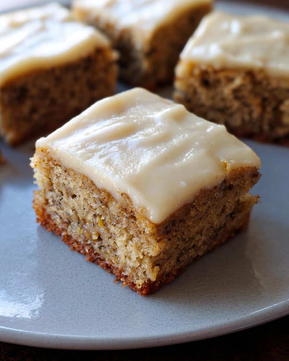 Close-up of a square Banana Bread Bars piece topped with thick, creamy frosting on a grey plate.