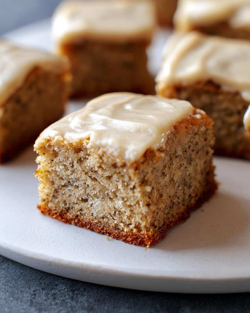 Close-up of a moist Banana Bread Bars square topped with light brown frosting on a white plate.