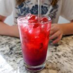A tall, sweating glass filled with a vibrant red Strawberry Acai Refresher, ice, and strawberry pieces.